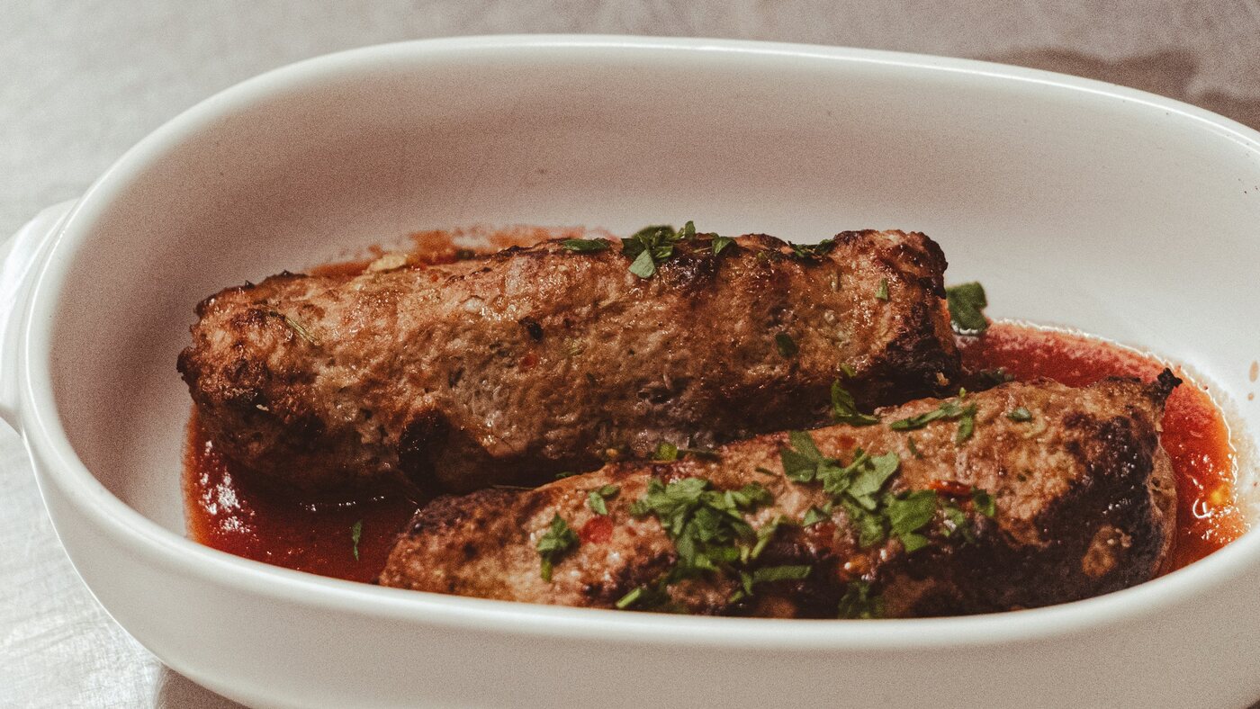 Dois bolos de carne assados em travessa branca, com molho de tomate e salsinha por cima