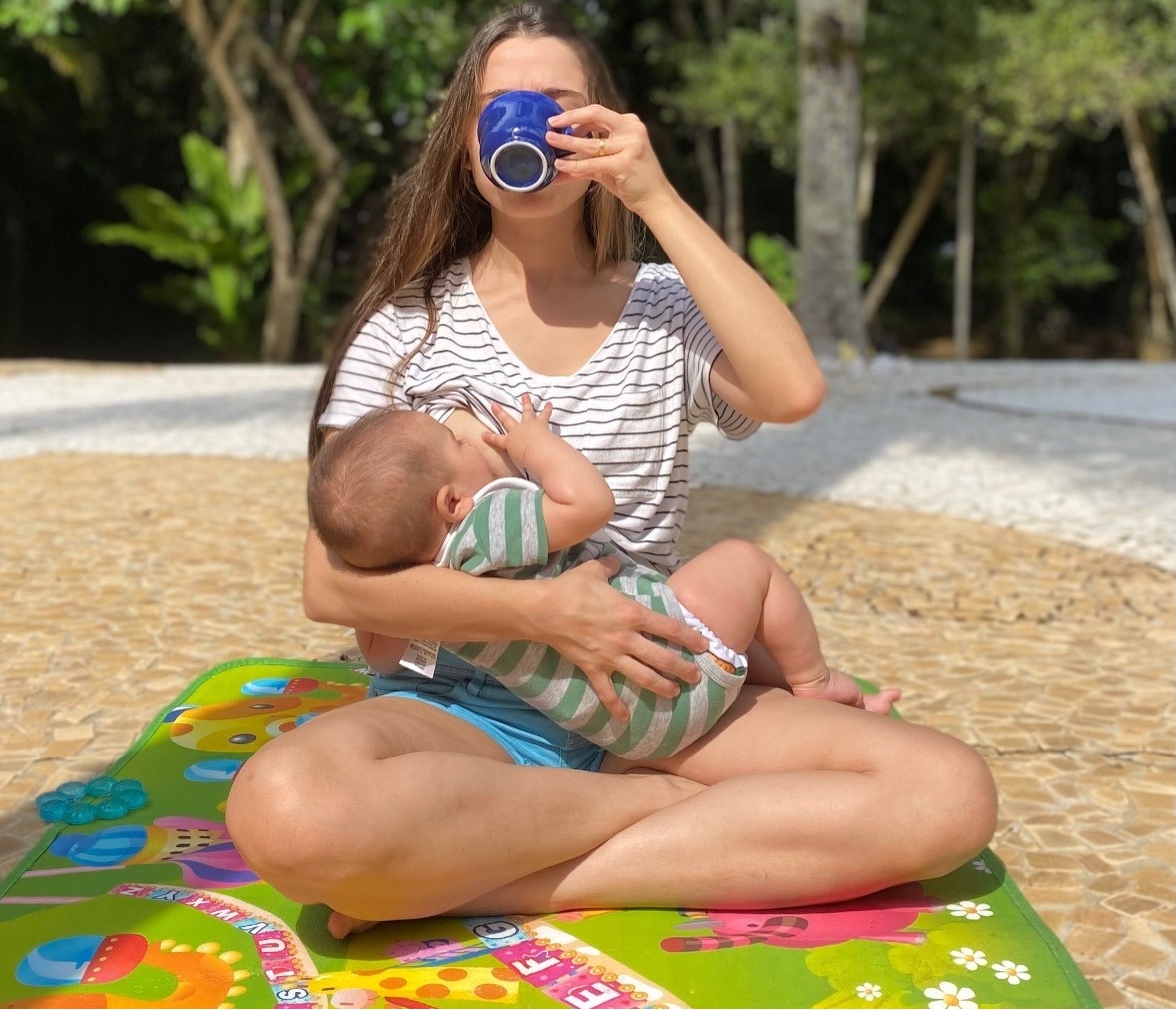 Mãe amamentando o bebê no colo, sentada em um tapete de atividades