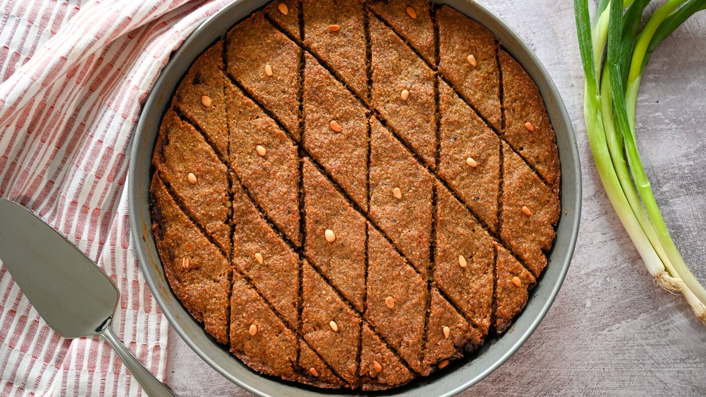 Quibe de forno assado em forma redonda, cortado em losangos e salpicado de castanhas